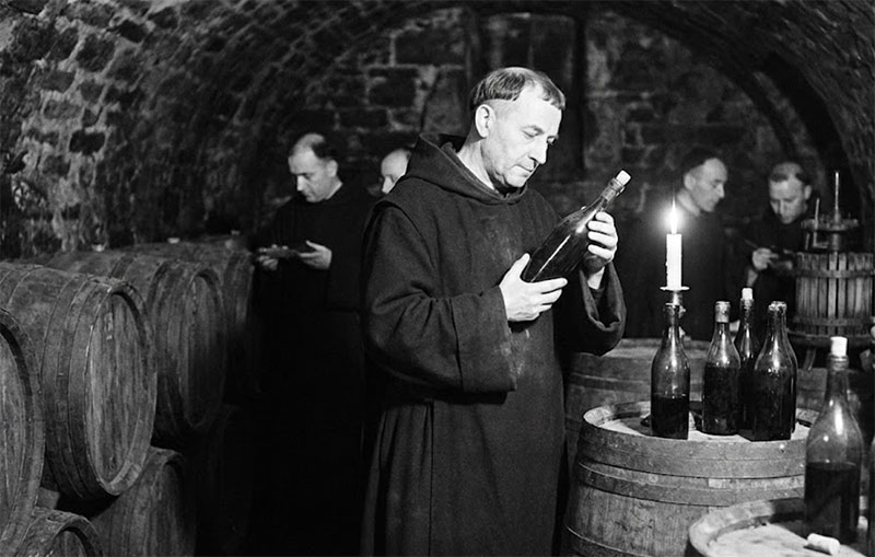 An artistic rendering of the Benedictine monk Dom Pérignon inspecting a bottle of wine in a candlelit cellar.