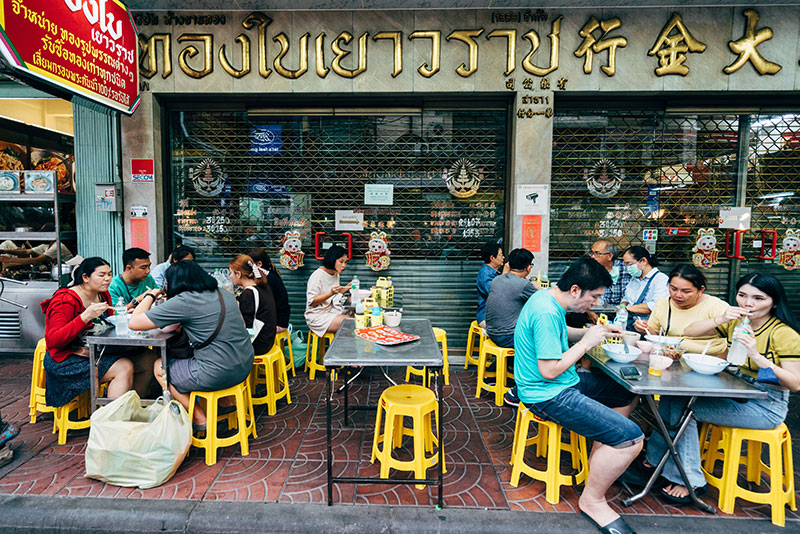 Locals and tourists eating street food at low tables with plastic stools on a busy street in Thailand at night.
