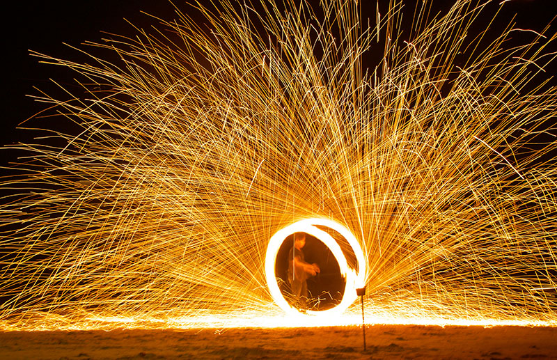 A fire spinner creating a circle of sparks on a dark beach in Thailand during a nightly fire show.