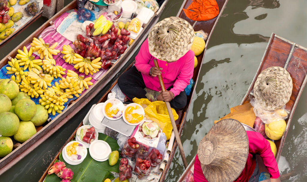 High-angle view of a vendor in a straw hat rowing a wooden boat filled with fresh fruit and flowers through a crowded canal at the Damnoen Saduak Floating Market