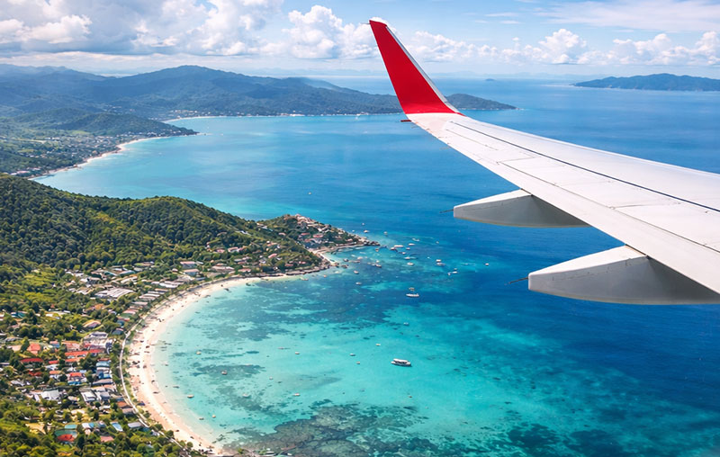 Aerial view of Koh Samui island from an airplane window, showing turquoise water, sandy beaches, and lush green coastline