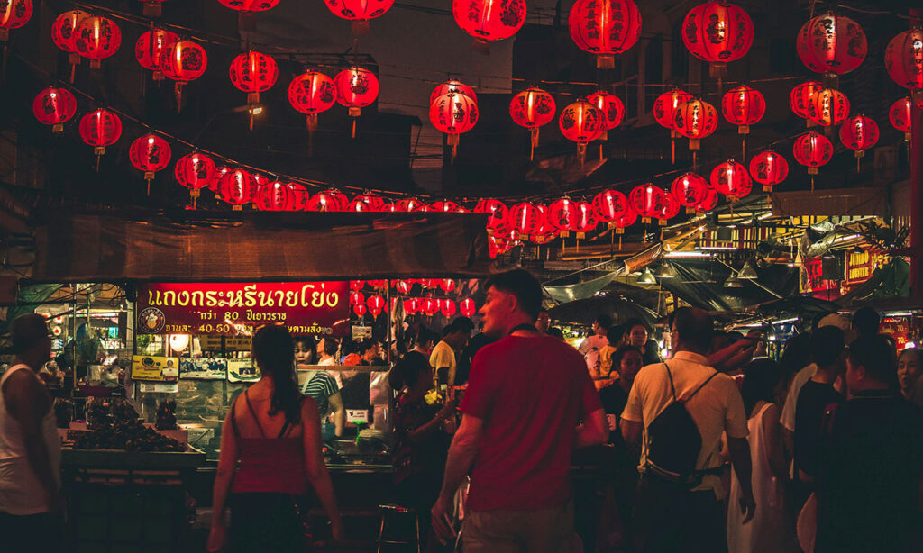 Night scene of a busy street food market in Bangkok's Chinatown (Yaowarat Road), with people dining at tables under rows of illuminated red lanterns.