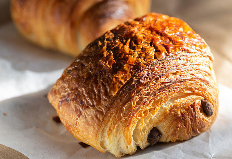 A close-up of a golden, flaky croissant and a pain au chocolat resting on bakery paper in the morning sunlight