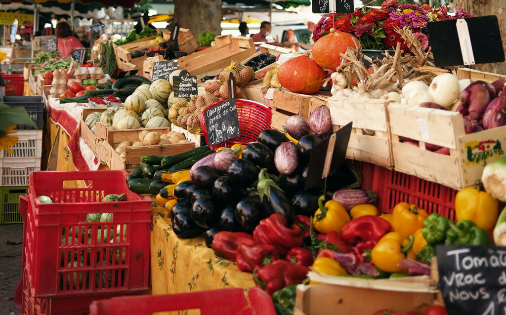 resh produce stall at an outdoor market in France, featuring colorful tomatoes, eggplants, and baskets.