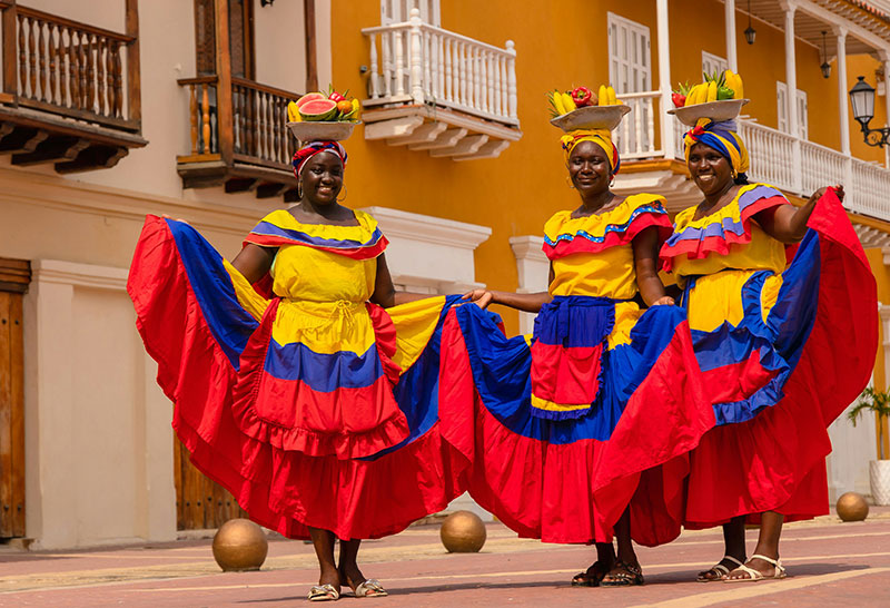 Two Colombian women in colorful traditional dresses and fruit headwear smiling in Cartagena, Colombia.