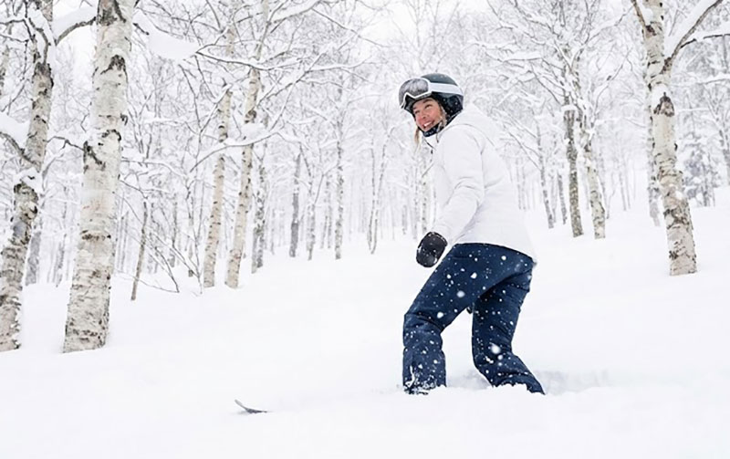 A female skier smiling while skiing through deep powder snow in a white birch forest in Niseko, Hokkaido.