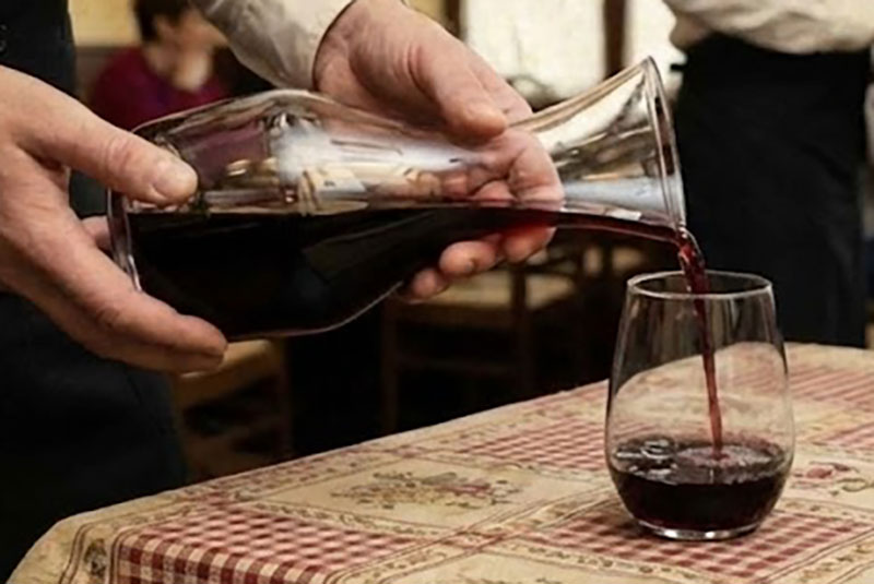 Waiter pouring red house wine from a glass carafe into a glass at a traditional Italian trattoria with a checkered tablecloth