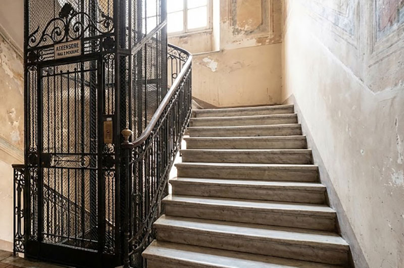 A tiny vintage elevator in an Italian building next to a steep marble staircase.