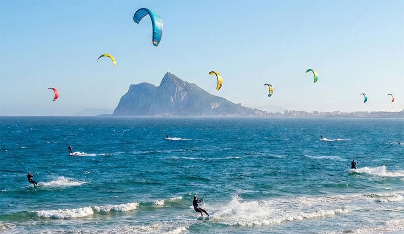 Dozens of colorful kitesurfers riding the wind on a sunny day at a beach in Tarifa, Spain, with the Rock of Gibraltar visible in the distance.