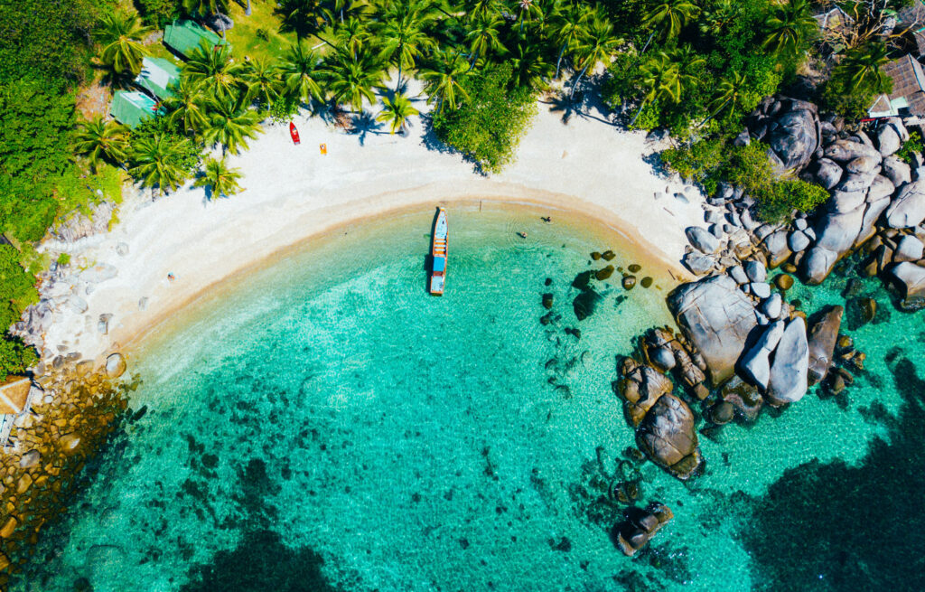 Top-down aerial view of a secluded sandy cove on Koh Tao with crystal clear emerald water and a single longtail boat.