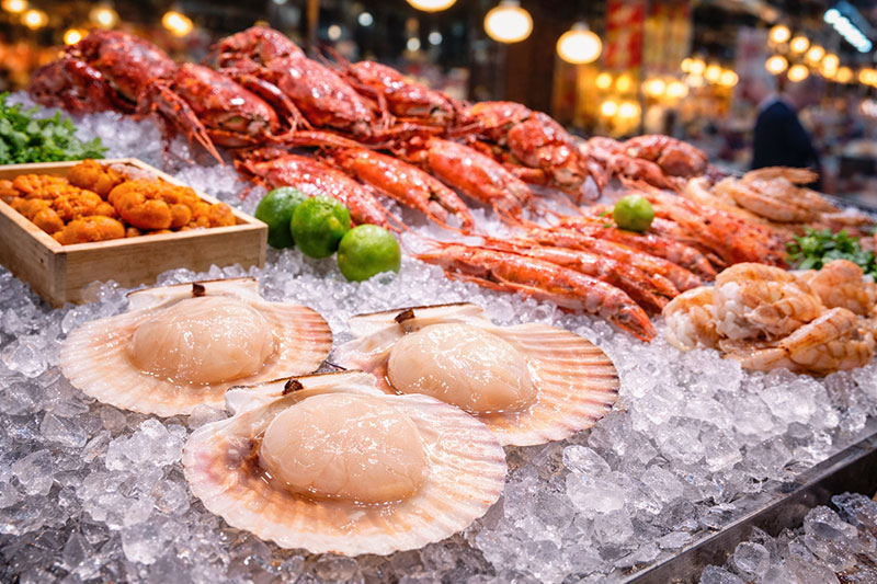 Raw giant scallops resting on their shells on crushed ice at Kuromon Market in Osaka, with fresh seafood displays blurred in the background.