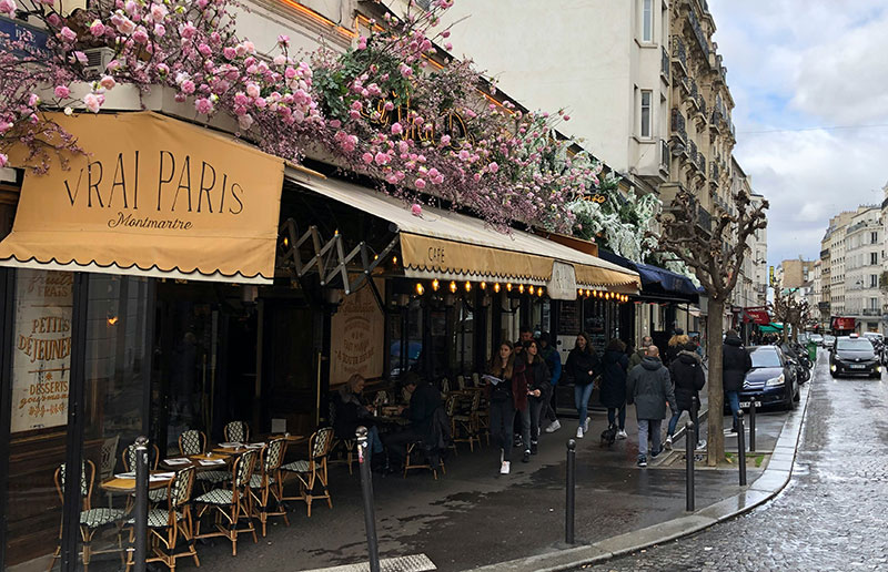 he exterior of Le Vrai Paris cafe on Rue des Abbesses in Montmartre, featuring a yellow awning, abundant pink flowers, and outdoor seating on a cobblestone street.