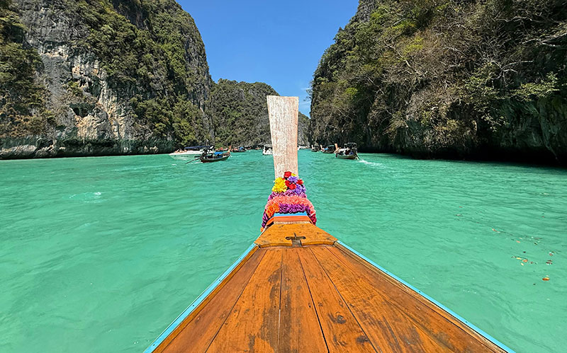 View from the wooden bow of a longtail boat decorated with colorful ribbons, heading toward the turquoise waters and limestone cliffs of the Phi Phi Islands.