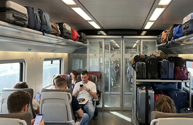 Interior of an Italian train showing suitcases stored in the overhead rack and larger bags in the rack at the end of the aisle.