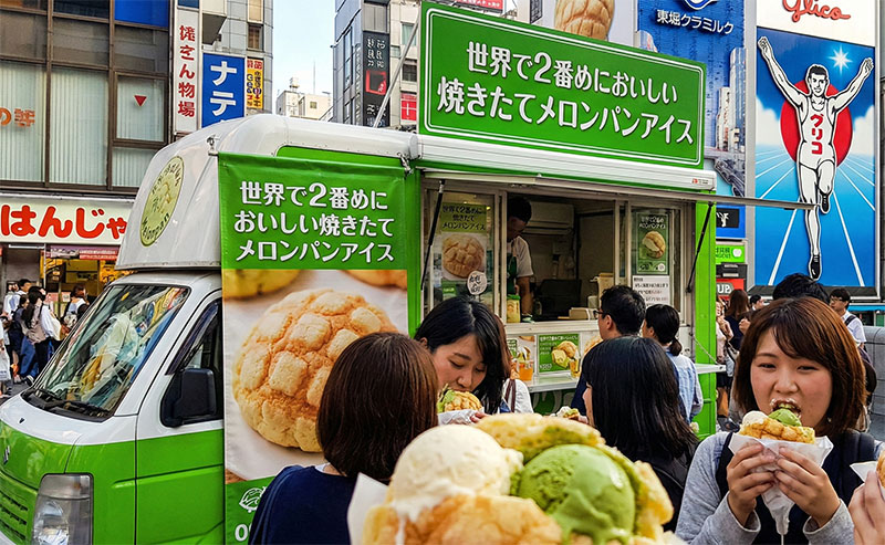 People enjoy warm melonpan stuffed with green tea and vanilla ice cream in front of the "World's Second Best Melonpan" food truck, parked in the bustling Dotonbori district of Osaka, with the Glico Man sign visible in the background.