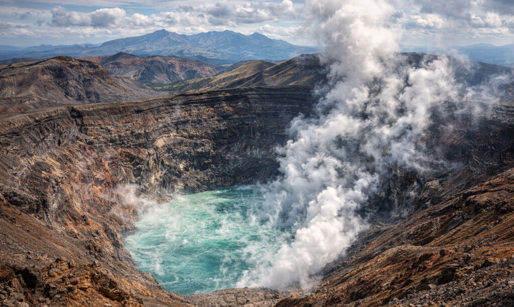 A breathtaking view of the massive Nakadake Crater at Mount Aso in Kyushu, Japan, featuring a steaming turquoise volcanic lake surrounded by rugged, rocky cliffs and distant mountains under a blue sky.