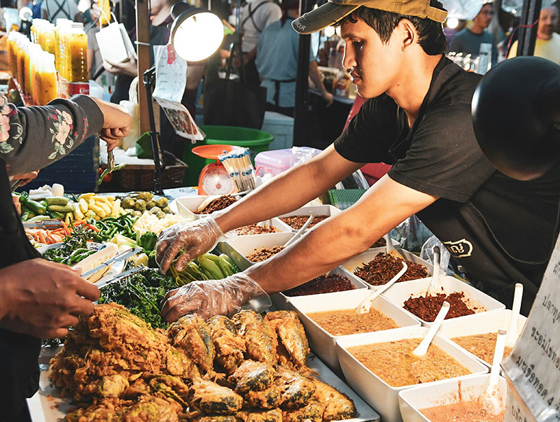 A street food vendor at a night market arranging trays of fried fish, fresh vegetables, and spicy dipping sauces (nam prik).