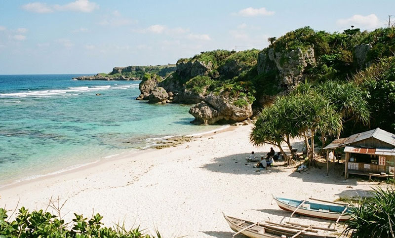 A pristine white sand beach in Okinawa featuring crystal clear turquoise water, lush greenery, and unique limestone rock formations along the coastline.
