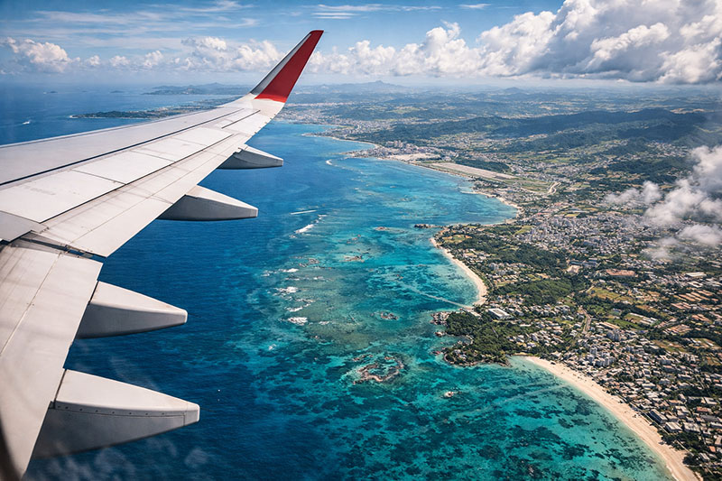 Aerial view of Okinawa Island from an airplane window during landing, showing turquoise ocean, coral reefs, coastline, and urban areas below.