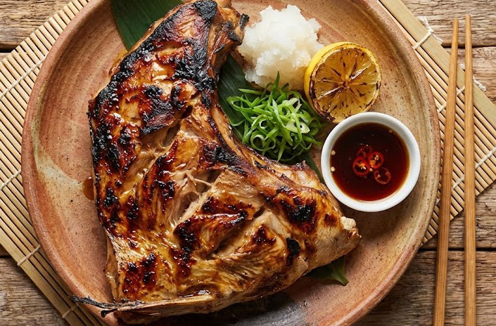 A top-down, overhead view of a grilled tuna collar (Maguro Kama) on a decorative plate, showing the charred texture, juicy meat, and side garnishes of lemon and grated daikon.