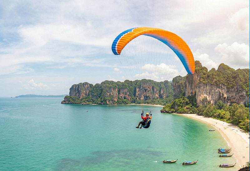 A paraglider pilot soaring high above the turquoise waters, longtail boats, and iconic limestone cliffs of Railay Beach in Krabi, Thailand.