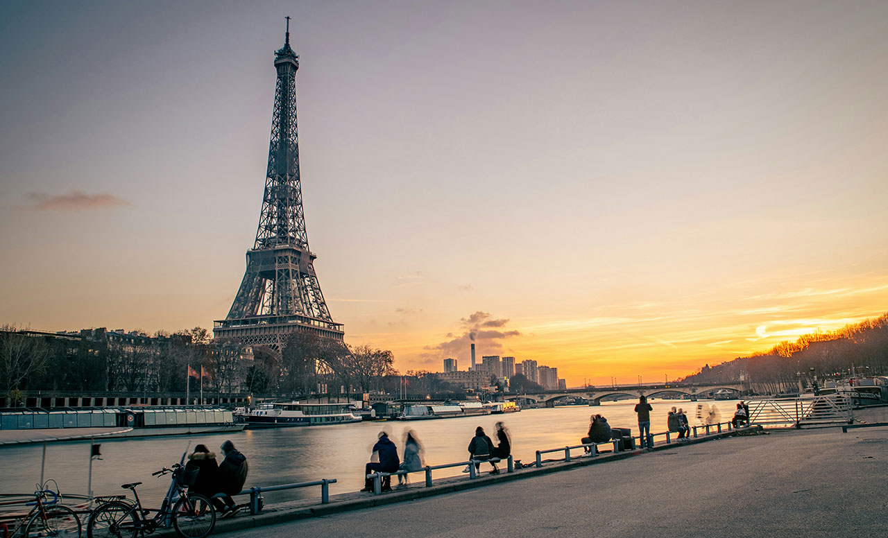 People walking along the Seine riverbank at sunset with the Eiffel Tower in the distance.