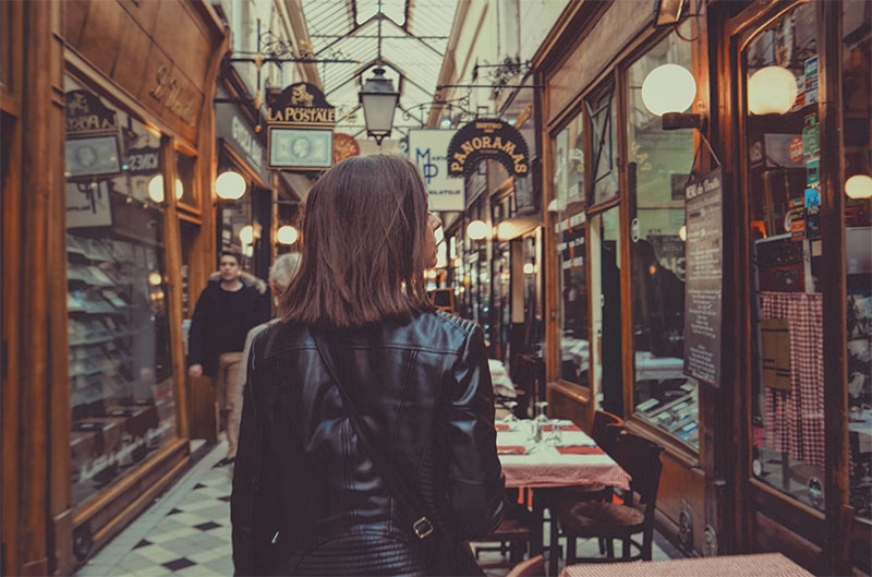A traveler walking through the Passage des Panoramas in Paris, a historic covered shopping arcade with glass ceilings, vintage signs like 'La Postale,' and checkered tablecloth restaurants.