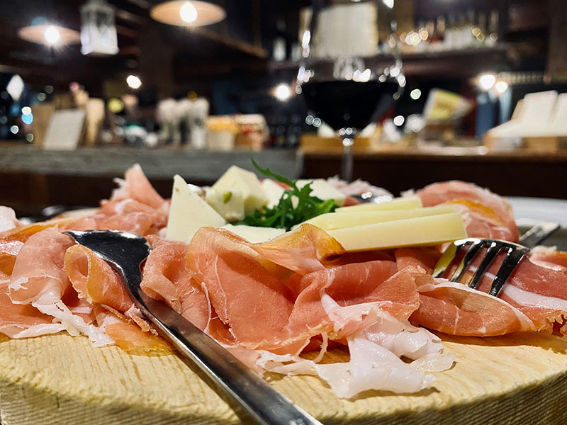 ooden board serving Prosciutto di Parma and parmesan cheese chunks with wine glasses in the background.