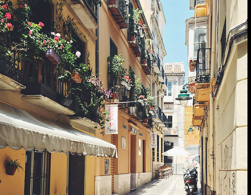 Narrow European street with balconies full of flowers, looking down the alleyway with buildings on both sides and a scooter parked.