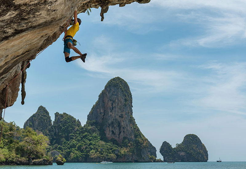A rock climber hanging from a limestone overhang at Tonsai Beach with a stunning view of the ocean and karsts in the background.