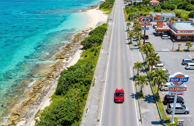 erial view of a car driving north on Route 58 in Okinawa, with the turquoise East China Sea on the left and retro American-style buildings on the right.