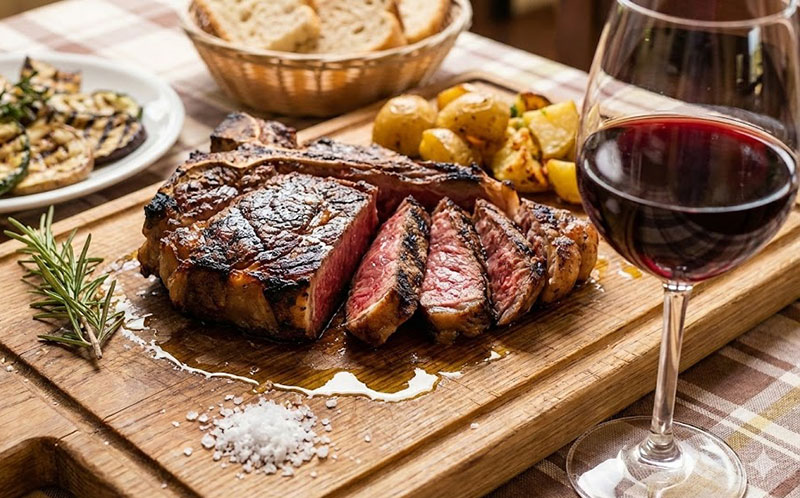 A thick-cut, grilled steak served on a wooden board with a glass of red wine, roasted potatoes, grilled zucchini, and a rosemary sprig.