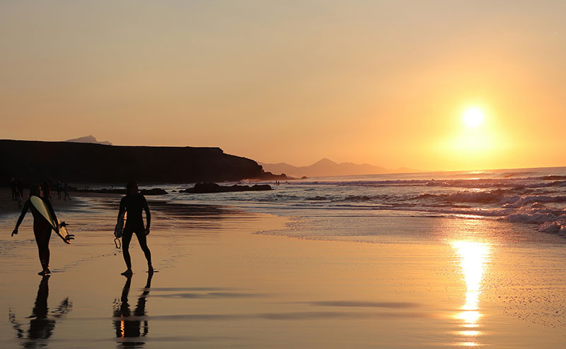 Two surfers with surfboards walk towards the ocean on a Canary Islands beach during a beautiful sunset.