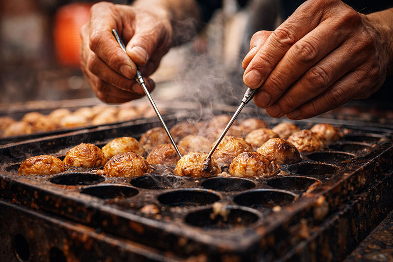 Close-up of a Japanese street food vendor’s hands using metal picks to flip golden-brown takoyaki on a hot cast iron griddle, with steam rising.