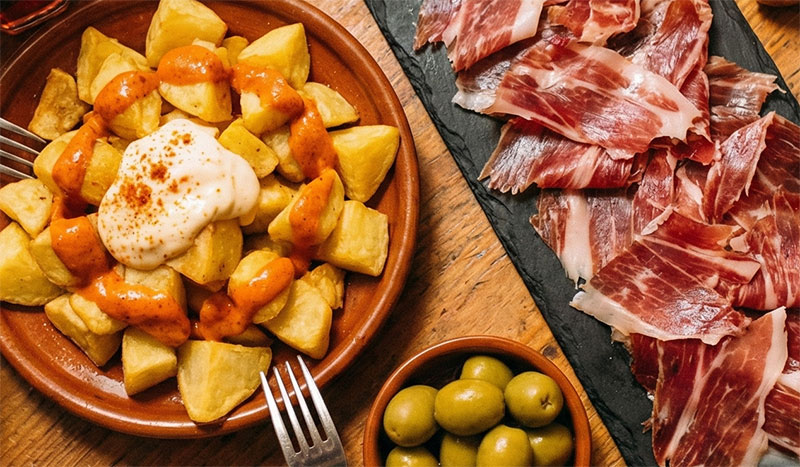 wooden table filled with plates of patatas bravas, jamón ibérico, olives