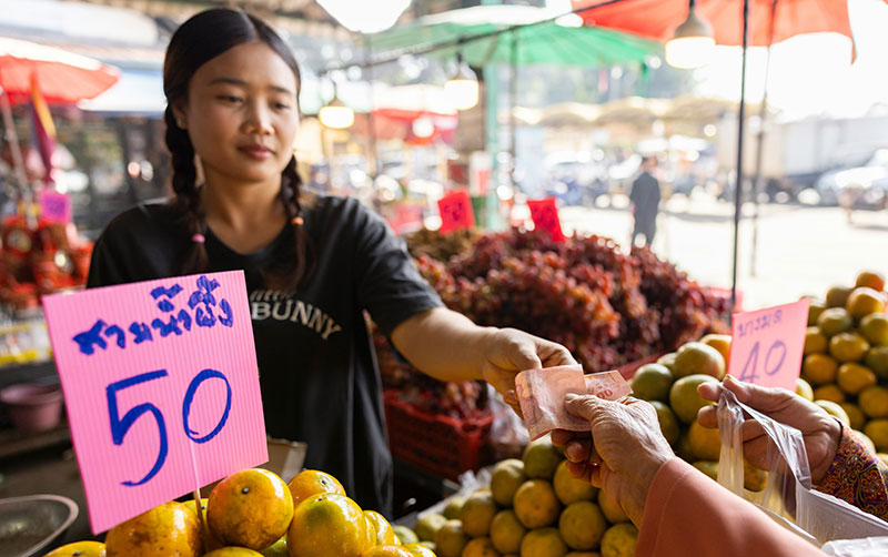 A close-up of a customer paying a market vendor with a Thai Baht banknote at a fruit stall in a bustling market.