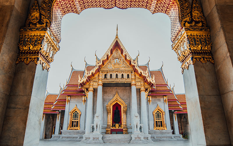 The ornate white marble main hall of Wat Benchamabophit (The Marble Temple) in Bangkok framed by a golden archway, illustrating a sacred space where modest dress is required.