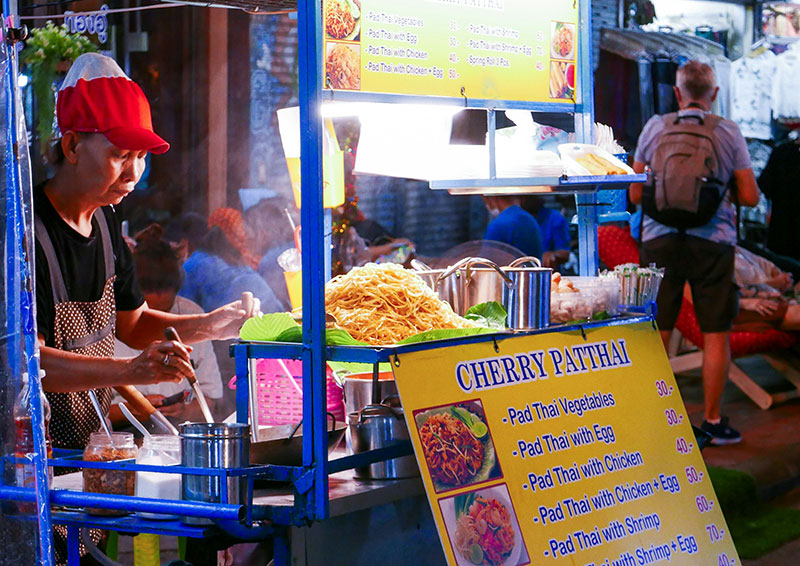 Thai street food vendor selling Pad Thai at a night market stall with fresh ingredients visible on the cart.