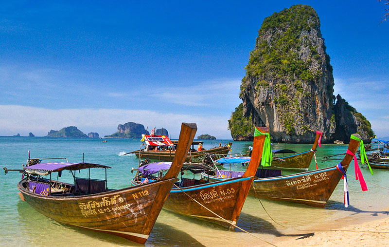 A traditional wooden longtail boat floating on turquoise water in front of a limestone cliff in Krabi, Thailand.