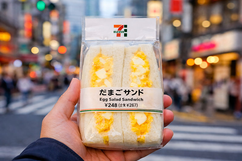 Close-up of a hand holding a famous 7-Eleven egg salad sandwich on a busy Tokyo street
