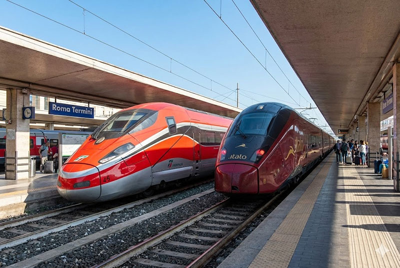 A red Trenitalia Frecciarossa train and a dark red Italo train parked side-by-side at a sunny Italian train platform.