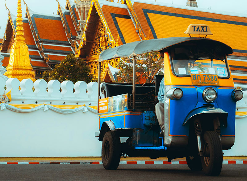 A blue and yellow tuk-tuk driving past the white walls and golden spires of the Grand Palace in Bangkok, a common location for tourist scams.