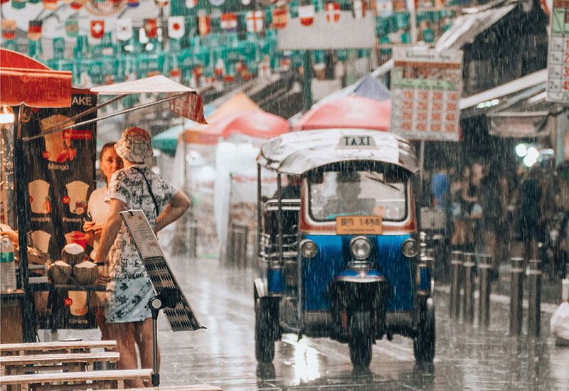 A blue tuk-tuk driving through a sudden torrential downpour on a busy Bangkok street, with rain blurring the colorful shop signs in the background.