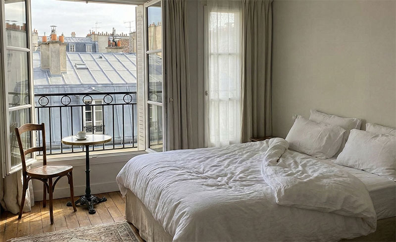 A bright but compact boutique hotel room in Paris with open windows facing typical zinc rooftops.