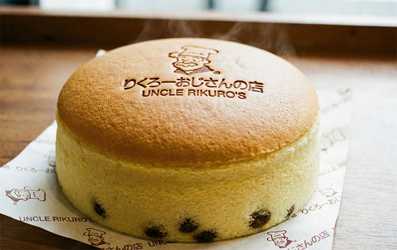 Close-up of a whole Uncle Rikuro's Japanese cheesecake showing the golden-brown top with the branded chef logo, fluffy souffle texture, and raisins visible at the base.