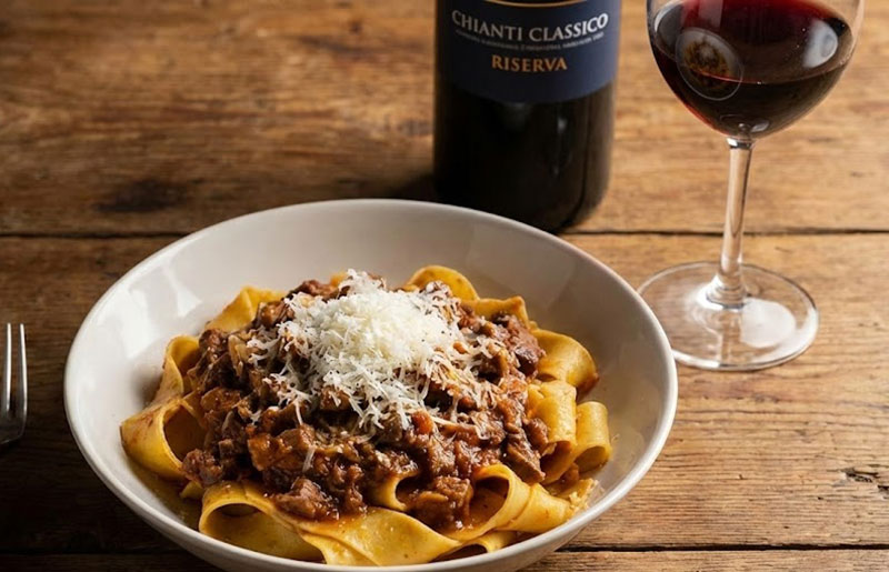 Plate of pappardelle pasta with wild boar ragù and grated cheese, paired with a bottle of Chianti Classico Riserva red wine on a wooden table