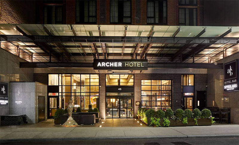 The modern exterior entrance of the Archer Hotel New York at night, featuring warm industrial-style lighting and "Archer Hotel" signage in Midtown Manhattan.