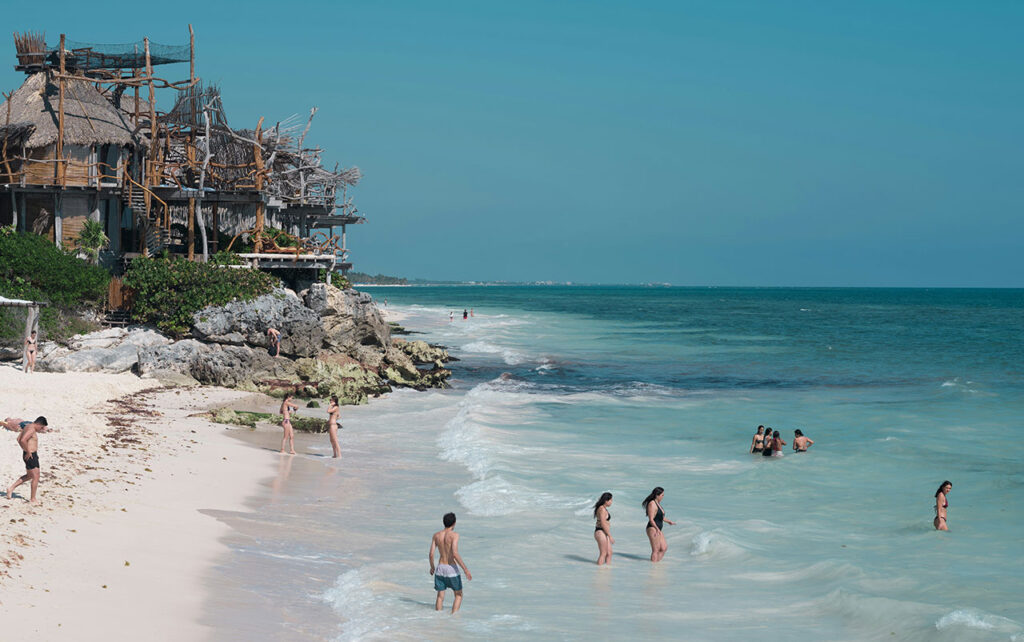 Scenic view of the white sand beach and turquoise water at Azulik Resort in Tulum, featuring rustic wooden architecture and lounge chairs.