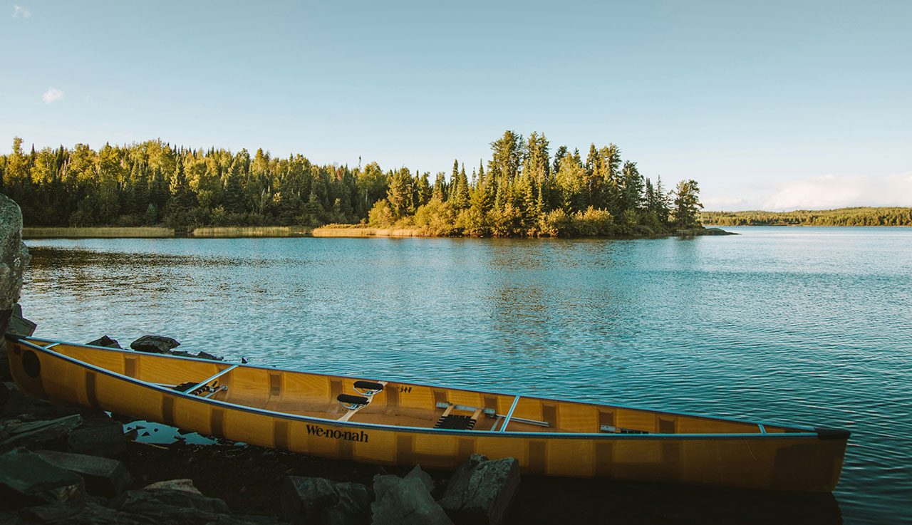 A tranquil view of a lake in the Boundary Waters Canoe Area Wilderness of northern Minnesota, featuring a yellow canoe resting on the shore of a pine-covered island under a clear blue sky.