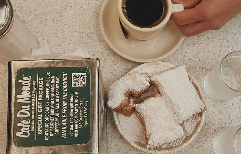 A close-up, top-down view of three fresh beignets heavily dusted with powdered sugar on a white plate, served with a cup of cafe au lait at Cafe Du Monde.